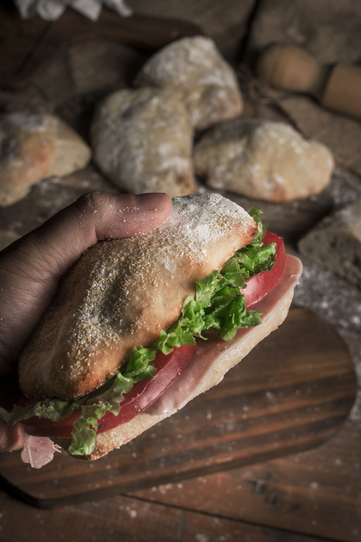 Ciabatta roll sandwich filled with tomato, salad leaves and ham, held in front of rustic homemade ciabatta rolls.